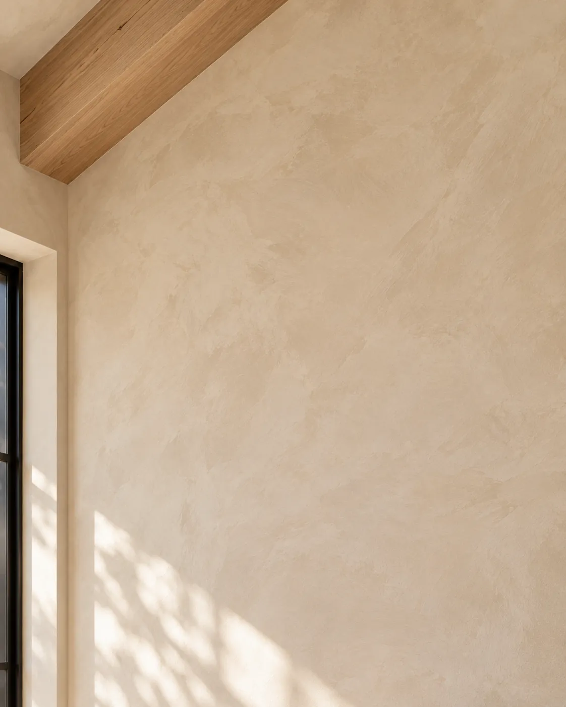 Close-up of warm stone beige limewash beside a black window frame and an oak beam, with soft brushed clouds and window light on the flat surface