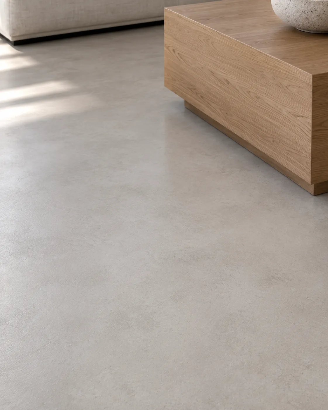 Close-up of a light cement gray microcement floor beside an oak coffee table, with soft cloudy trowel movement and daylight across the matte surface