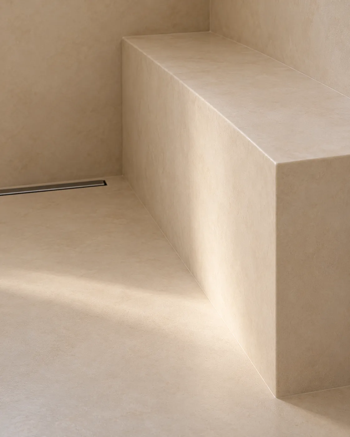 Close-up of a sand microcement shower bench meeting the floor beside a linear drain, one continuous matte surface with wrapped edges