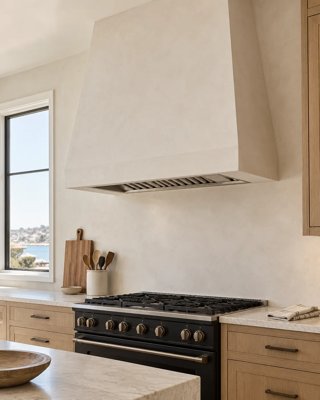 Kitchen with a warm white microcement range hood and wall behind a black range, white oak cabinets, a stone island and a window to the harbor