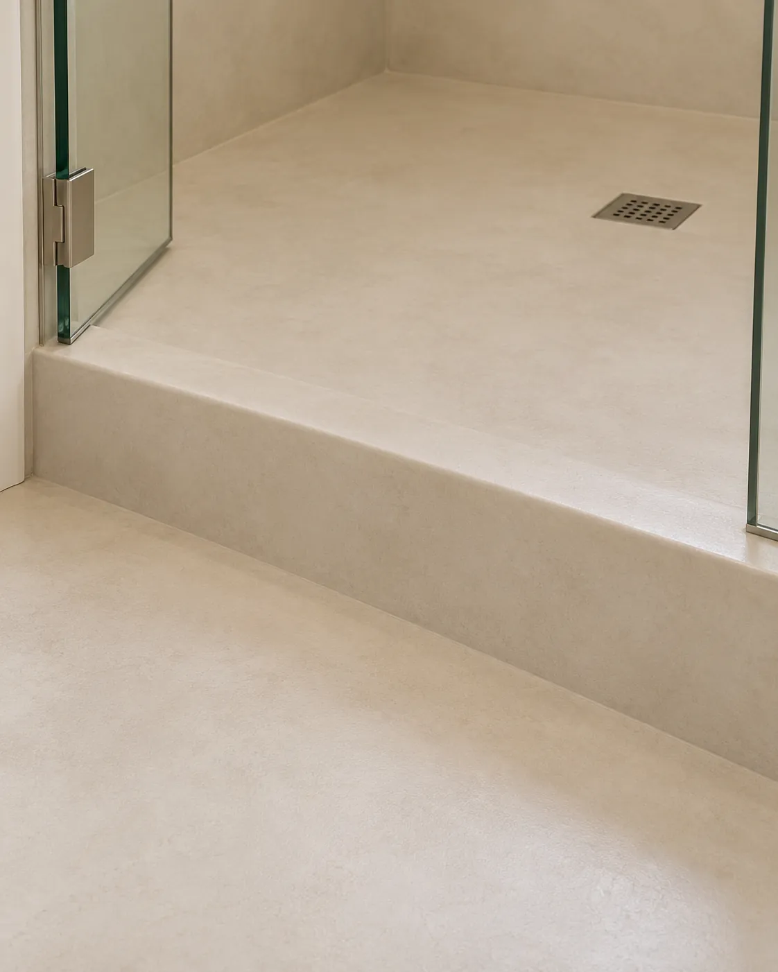 Close-up of a warm white microcement shower curb and floor beside a glass door hinge, with a square drain and softly rounded wrapped edges