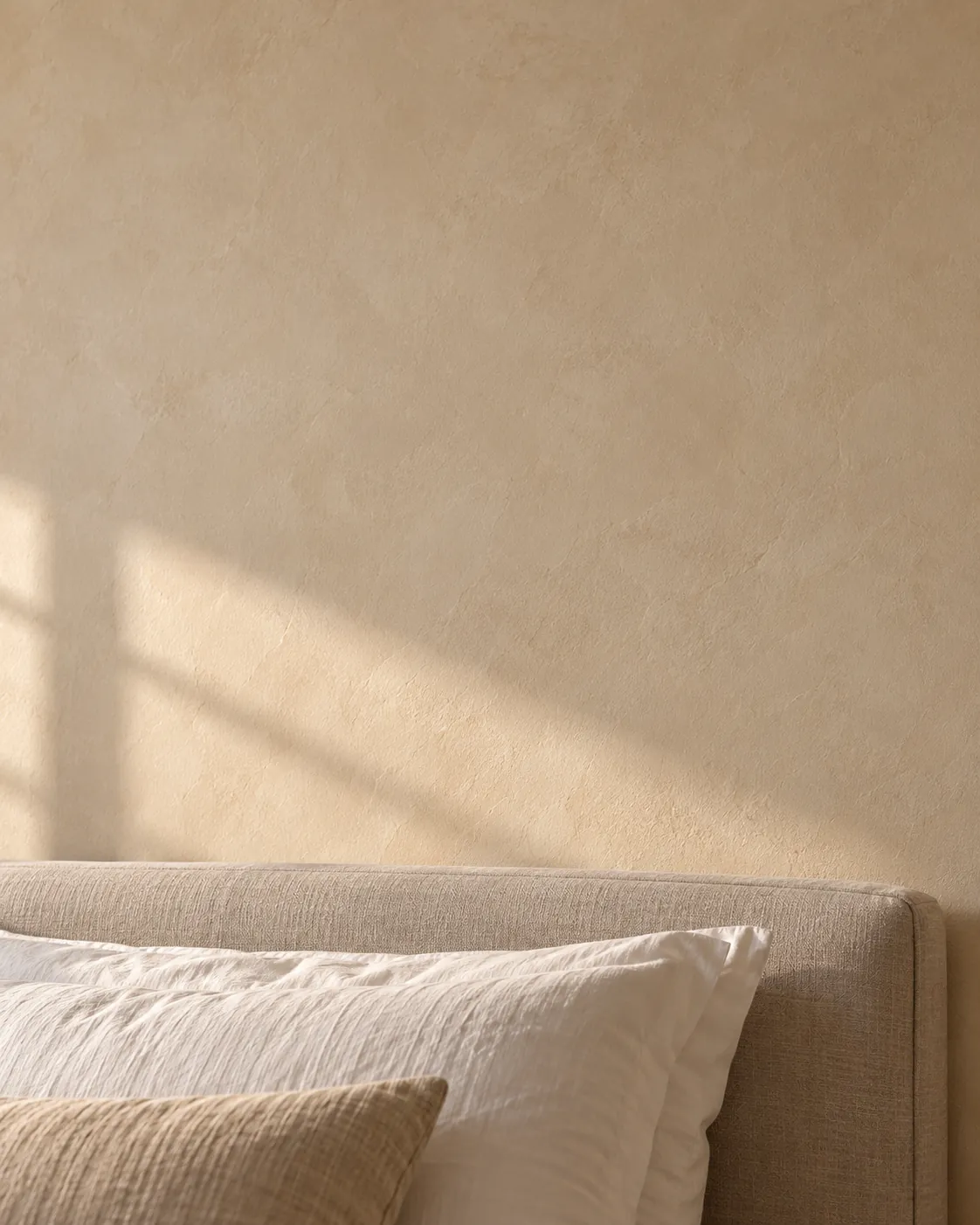 Close-up of oatmeal Roman clay above a linen headboard, with soft sand-colored clouds and window light falling across the matte wall
