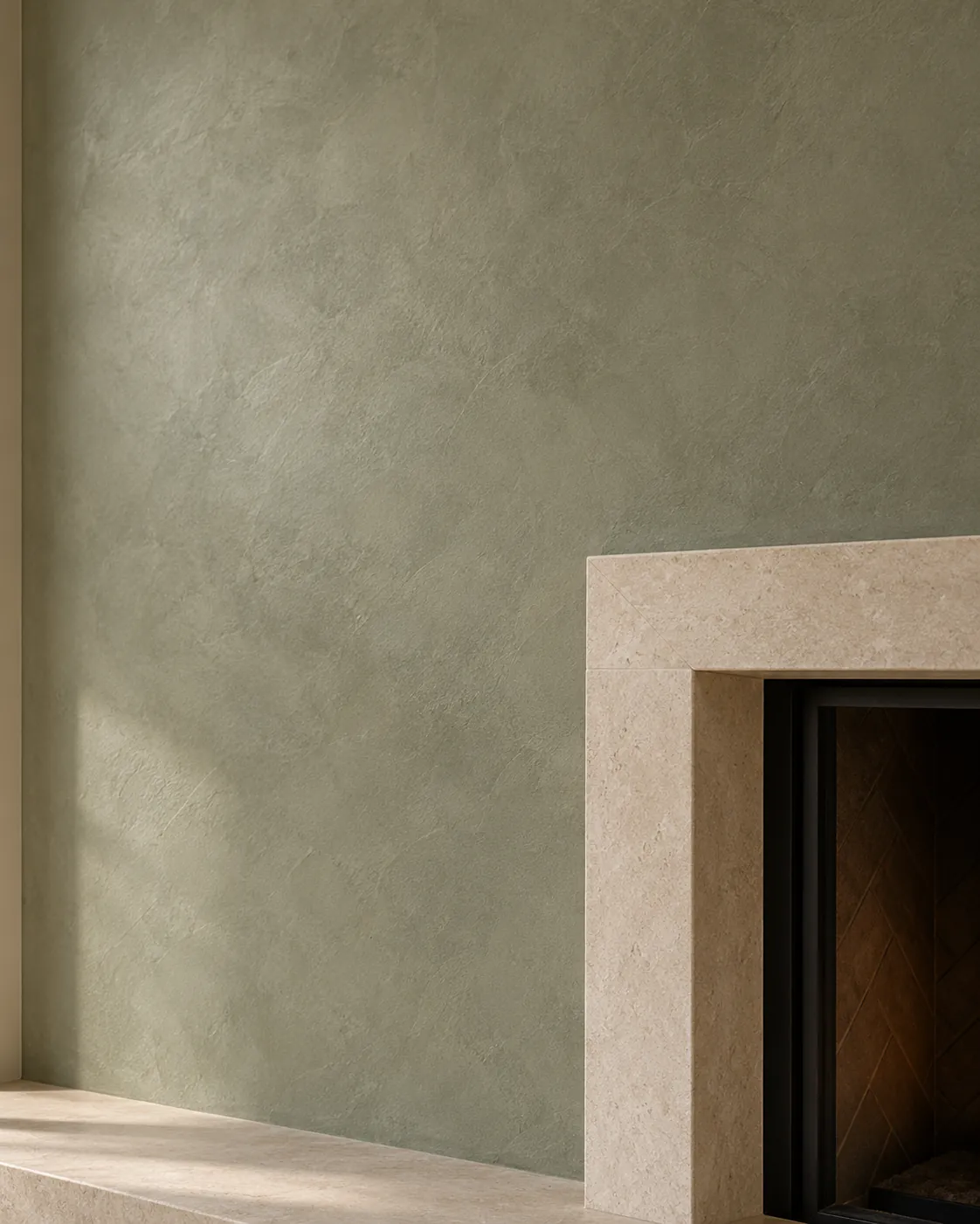 Close-up of sage green Roman clay beside a limestone fireplace surround, with soft cloudy shifts and late daylight on the wall