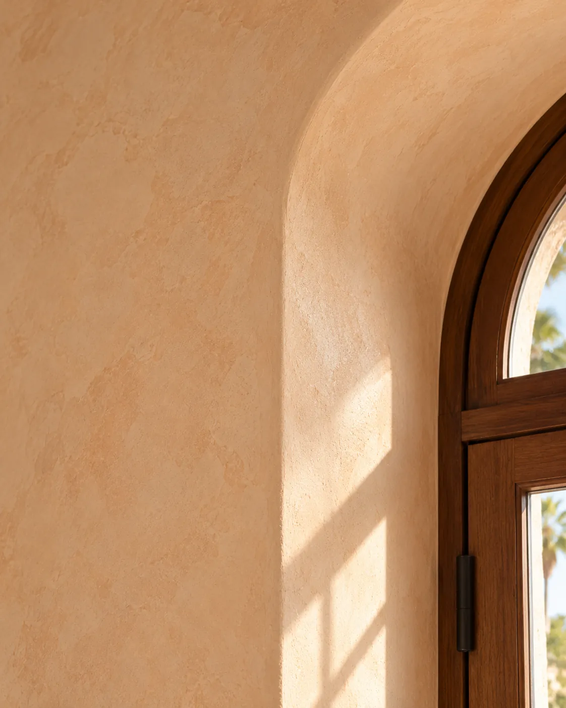 Close-up of soft peach Marmorino plaster wrapping the rounded reveal of an arched wood window, with morning light across the matte surface