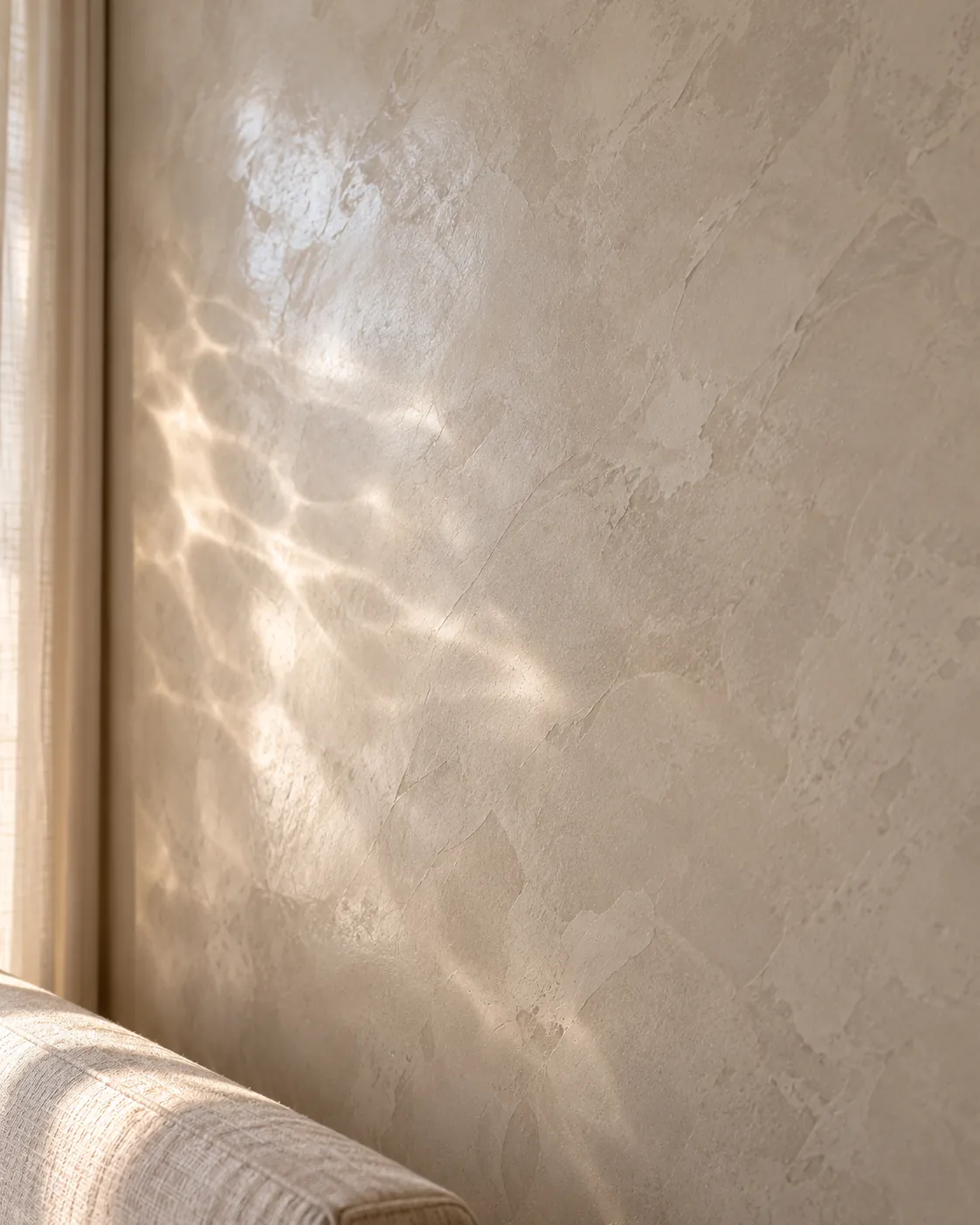 Close-up of warm gray polished Venetian plaster beside a linen curtain, with rippled light reflected from water moving across the satin surface